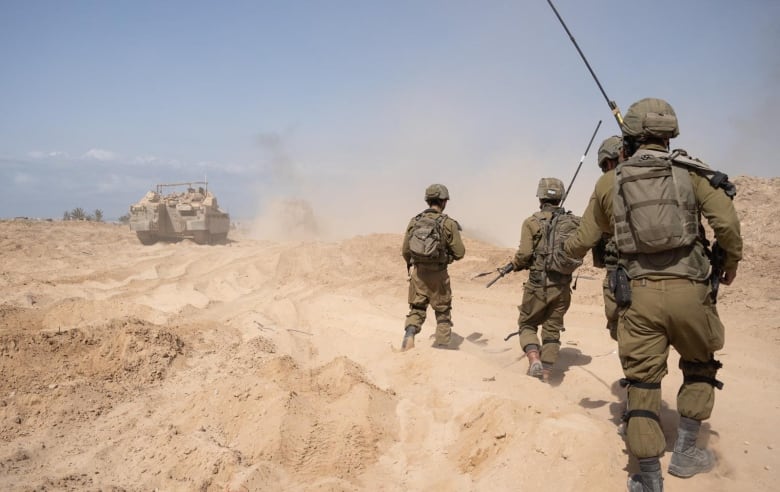 Four soldiers march across a desert lanscape -- with an armoured vehicle leading the way in the middle distance. 