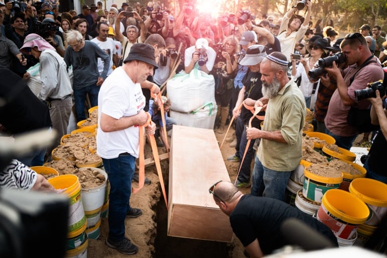 Pallbearer use ropes to lower a coffin into a grave as people gather and take photos around them.