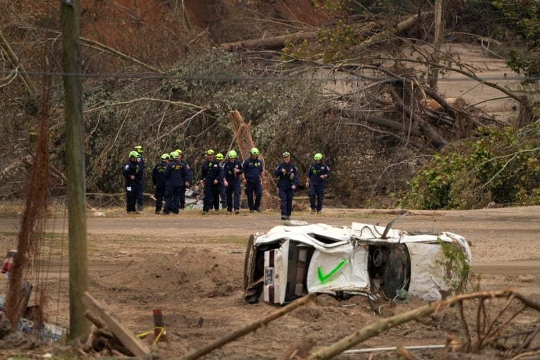 Search and rescue personnel walk on a muddy field with a car on its side in the foreground.