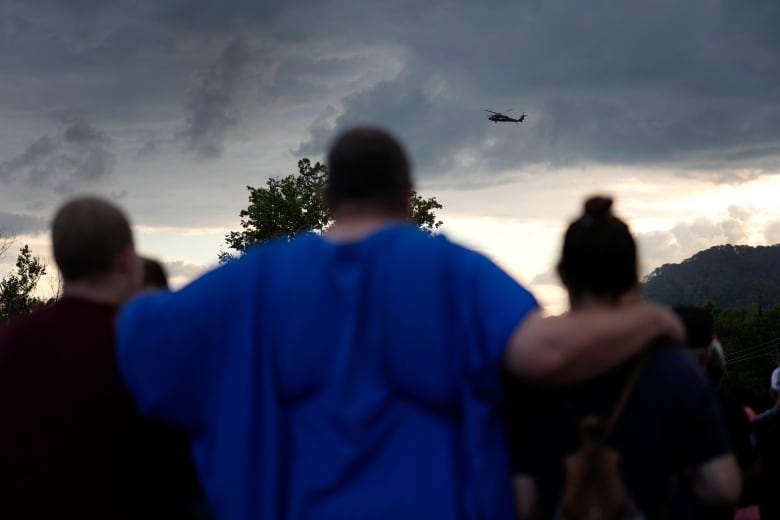 A person stands with their arms around two other people as a helicopter flies overhead.