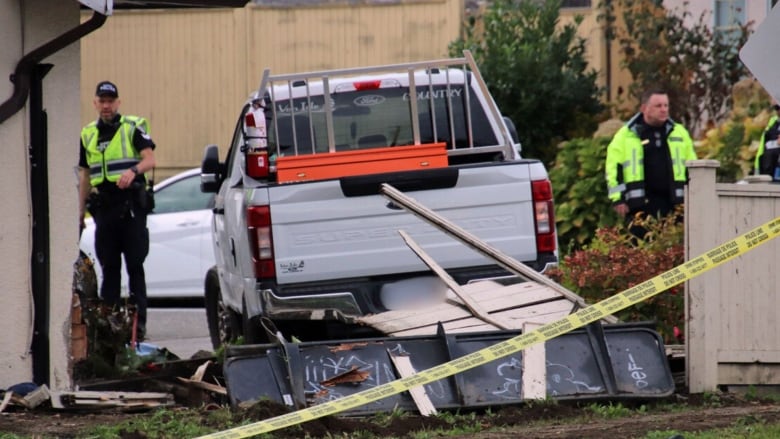 A pickup truck lies behind crime tape and a broken fence, as two police officers look on.