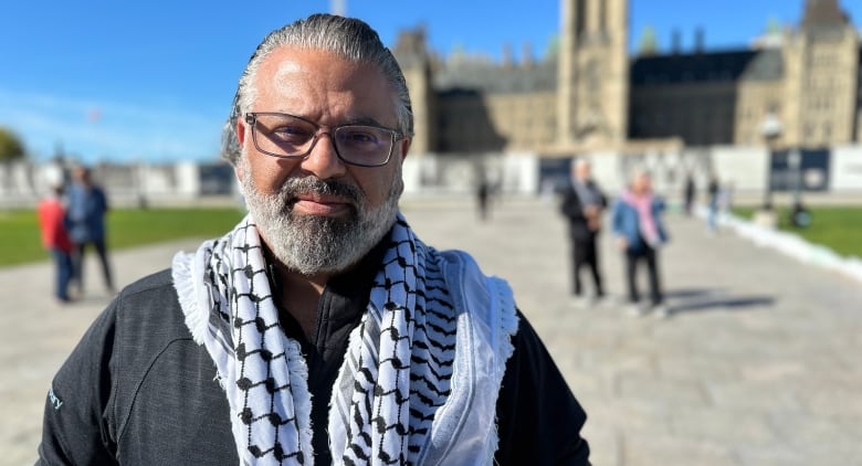 A man with glasses stands in front of Canada's parliament with a Keffiyeh draped around his shoulders.