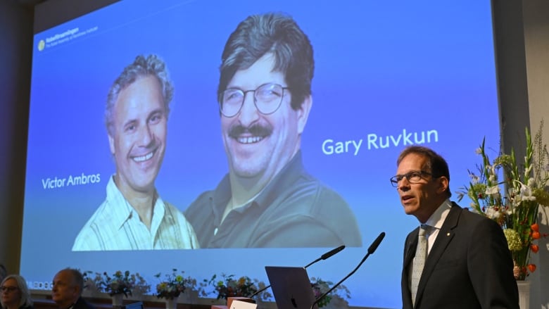 A cleanshaven bespectacled man speaks at a podium off to the side as a large projector displays the images of two men, one clean shaven and the other bespectacled with a mustache.