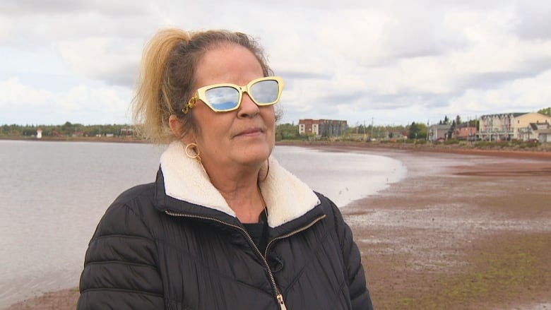 Woman with winter coat and sunglasses standing on shoreline.