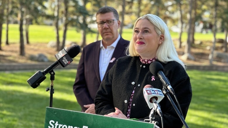 A woman wearing a colourful shirt and a black sweater stands behind a podium. A man looks on over the woman's right shoulder. 