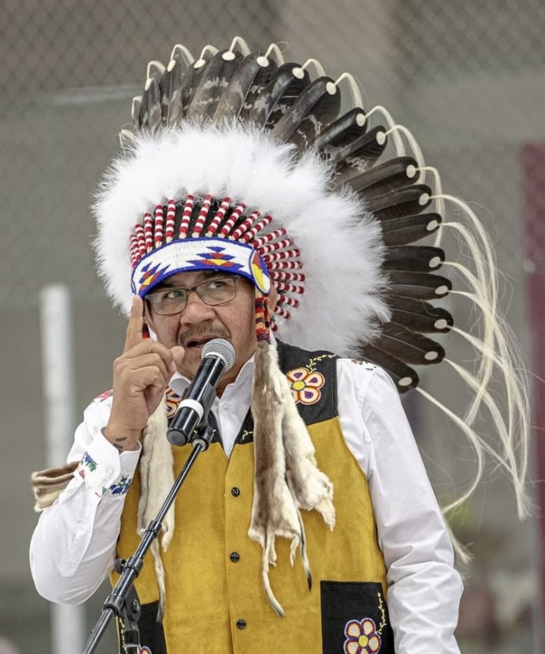 Man with glasses, and wearing a headdress is speaking into a microphone, holding his index finger up. 