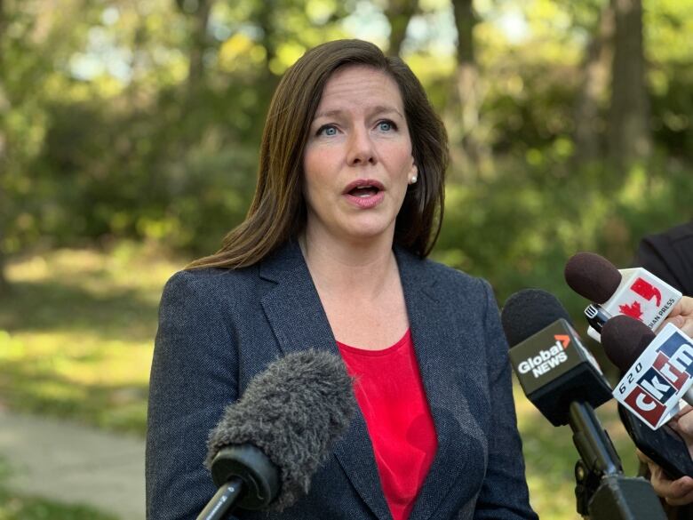 A woman speaks with microphones in front of her. The woman is wearing a red shirt and a dark-grey blazer.