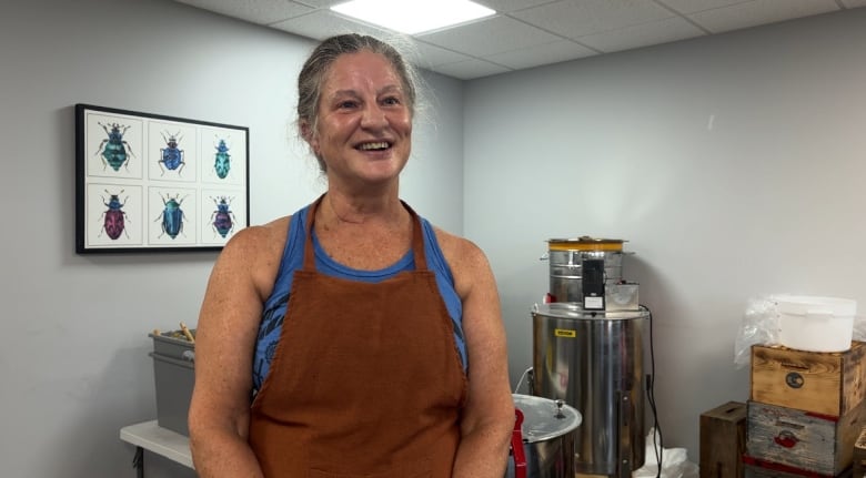 Woman with grey hair pulled back, smiling. She's wearing a blue sleeveless shirt with a brown apron. Behind her is a cylindrical metal can.