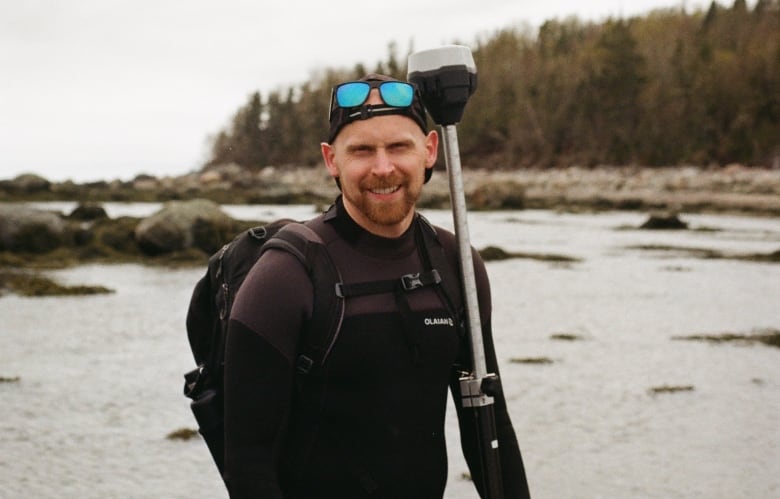 A man is standing on a beach wearing a wetsuit and backpack. 