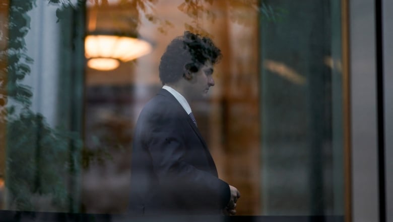 A man is seen in profile view through a window. Reflections of tree branches are visible on the window pane and the man is in a suit. 