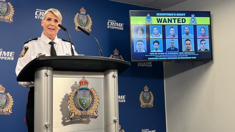 A police officer stands at a podium. A TV screen shows a list of offenders wanted by police. 