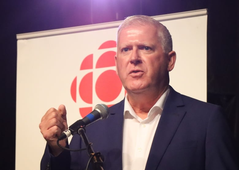 A man in a dark blue suit with a white shirt and no tie makes a fist as he speaks into a microphone.