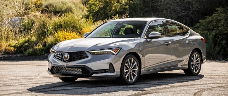 A silver car is parked in front of a green foliage background in an outside parking lot. 