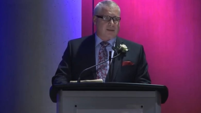 A man with grey hair, black glasses and a suit with a purple dress shirt stands at a podium.