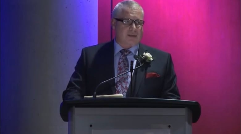 A man with grey hair, black glasses and a suit with a purple dress shirt stands at a podium.