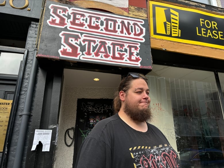 A bearded man stands in front of a sign that reads 