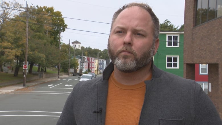 Man in grey jacket undone, wearing an orange sweater underneath. He's standing on the corner of a street.