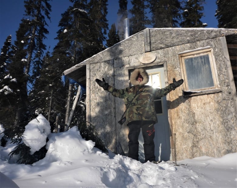 A woman wearing a camoflauge parka and snow pants stands in front of a large wooden hut in the snowy wilderness.