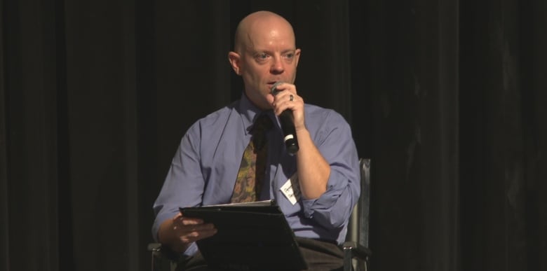 A man ina  shirt and tie sits on a darkened stage speaking into a microphone.