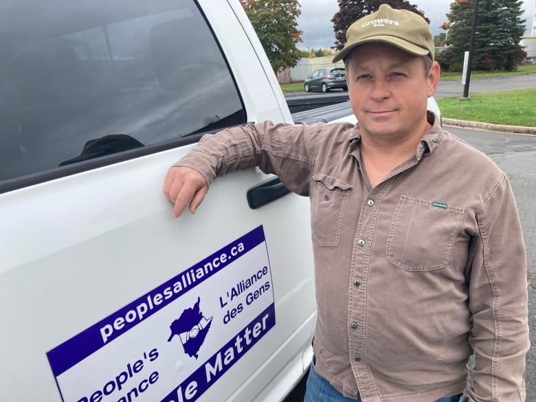 Aman in a beige ballcap and shirt leans on a white truck bearing the logo of the Alliance party.