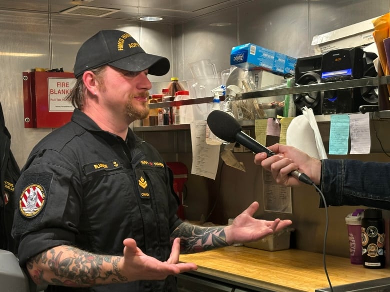 A middle aged man in a navy uniform speaks with a reporter in a navy ship's galley