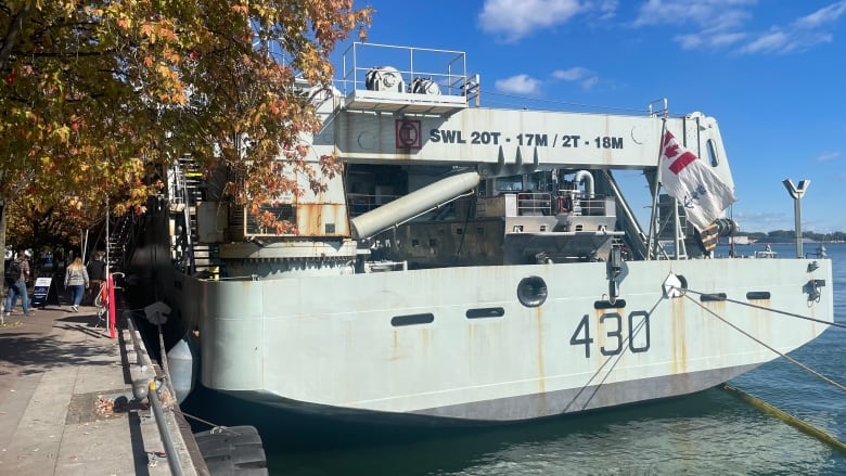 The stern of a large navy patrol ship is visible, docked in the water of TOronto harbour. The rest of the ship is obscured by trees on the shore. It's a sunny fall day