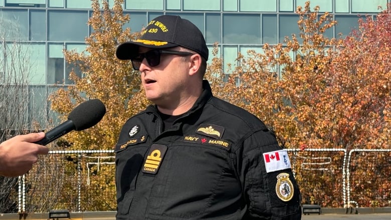 A naval commanding officer in uniform and sunglasses speaks to a reporter on a ship's deck. It's a sunny fall day. The ship is docked and autumn leaves are visible behind