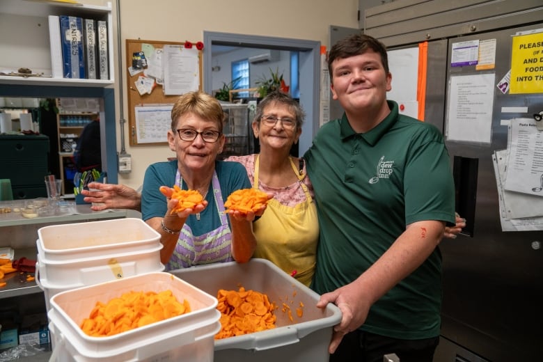 Three people stand in a kitchen around containers of carrots.