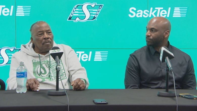 Two men are sitting at a black table. The man on the right is Black and wearing a black button up shit. The man on the left is Black and wearing a grey hoody, he is an older man. They are in front of a green background that has the Saskatchewan Roughriders logo. 