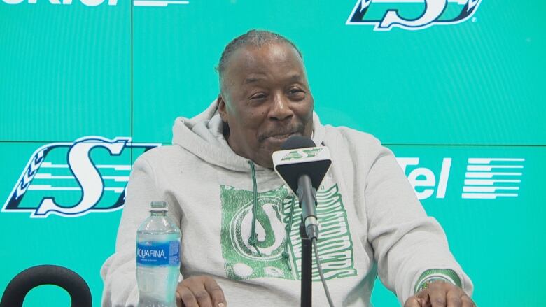 An elderly Black man wearing a grey hoody sits behind a microphone in front of a green screen with the Saskatchewan Roughrider logo on it. 