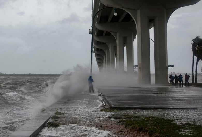 A reporter in rain gear is splashed by waves while a group of people stand around a video camera some distance away, recording the wind and rain hitting a causeway in Florida. 