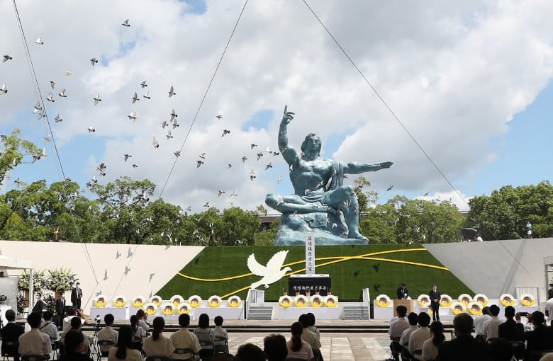 Doves fly near a large statue of a human figure, with dozens of people seated in what appears to be an outdoor ceremony.