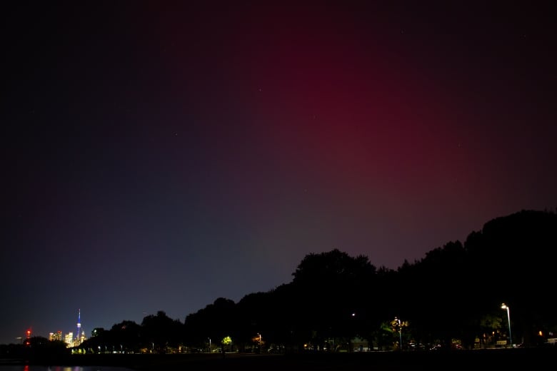 The CN Tower and downtown Toronto are visible in the distance at night. In the foreground, the silhouette of a beach and trees. In the sky, the faint red of the Northern Lights