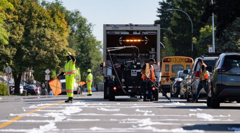 Photo shows a street where the pavement is covered in stripes of something white, perhaps related to fixing the road. In the distance, a truck sits with its back to the camera, and workers stand around. One holds a long device that points at the pavement. 