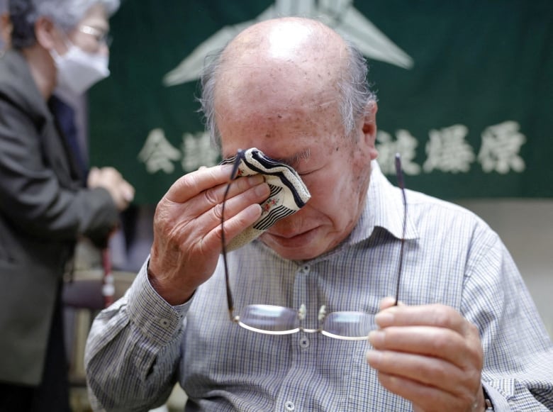 An elderly, balding Asian man holds his glasses with his left hand while dabbing his eyes with a handkerchief in his right hand.