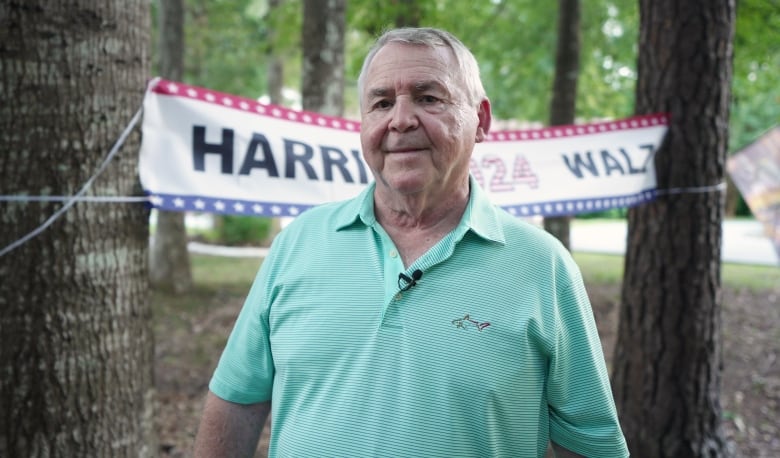 A man with grey hair, wearing a light green golf shirt, stands between some trees.