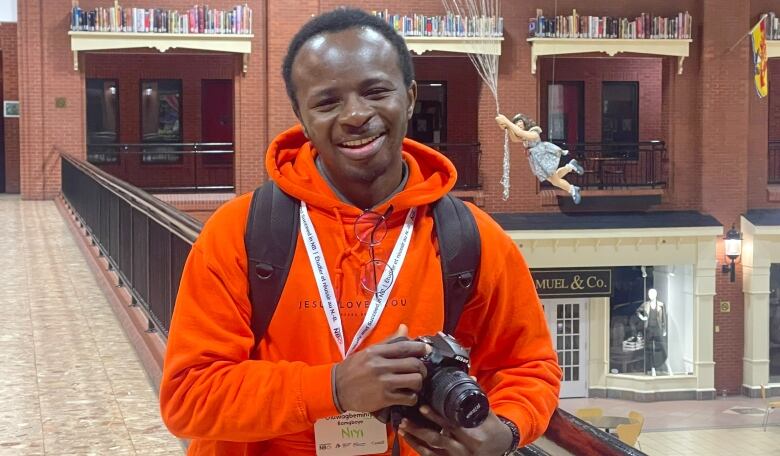 A smiling man, wearing a neon orange hoodie, standing against a railing and holding a DSLR camera. 