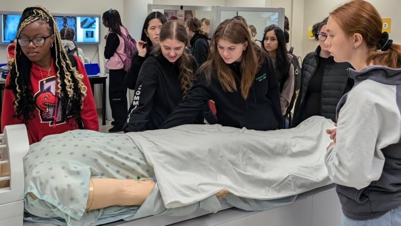 Five young women in a laboratory surround a dummy lying on gurney as a simulated patient.