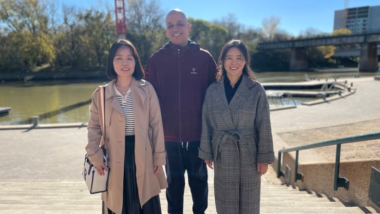 Three people smile as they stand in front of a river in a park space.