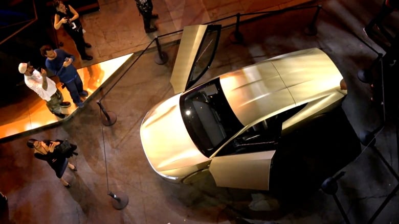 A car with its gull-wing doors open is seen from above. Several people stand around taking photos of it. 