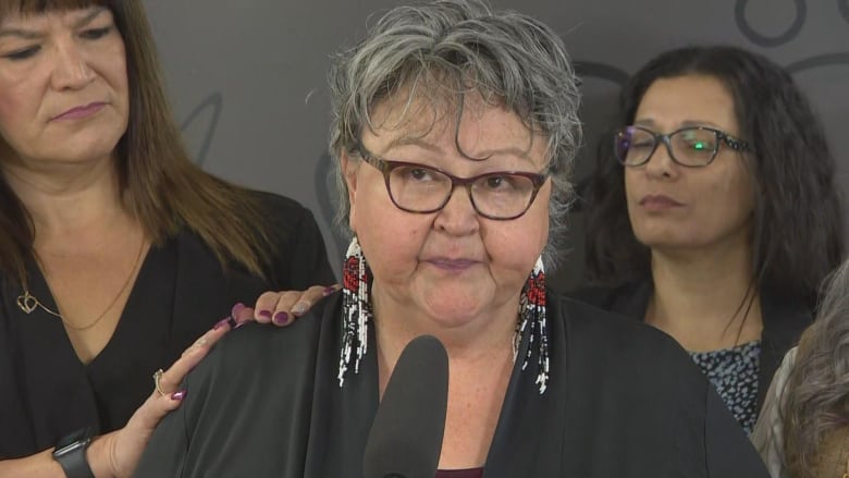 A woman in a black blouse stands behind a microphone, while another woman's hands rest on her right shoulder. 