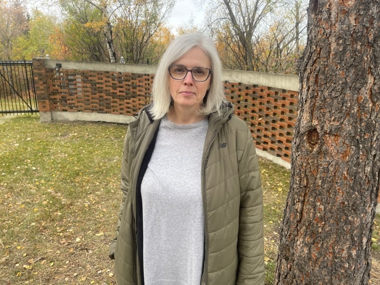 A woman with grey hair stands outdoors in autumn wearing a green jacket, with fall foliage behind her.