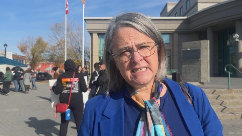 A woman in wire-rimmed glasses and grey hair in a blue suit with a multi-coloured scarf stands outside a municipal building. 