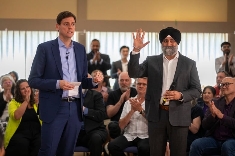 A tall man in a blue suit stands next to a man wearing a turban and a suit in front of a crowd of people. 