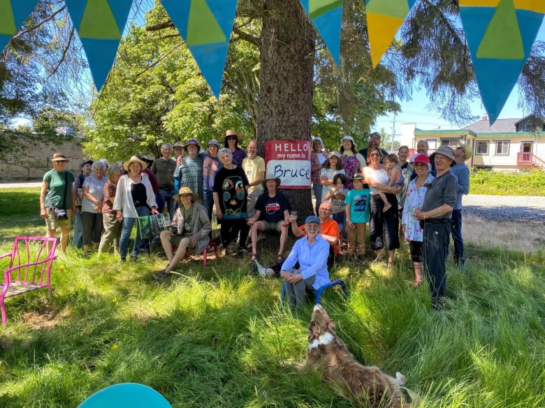 A group of people gather around a large tree, which has a sign on it reading 'Bruce.'