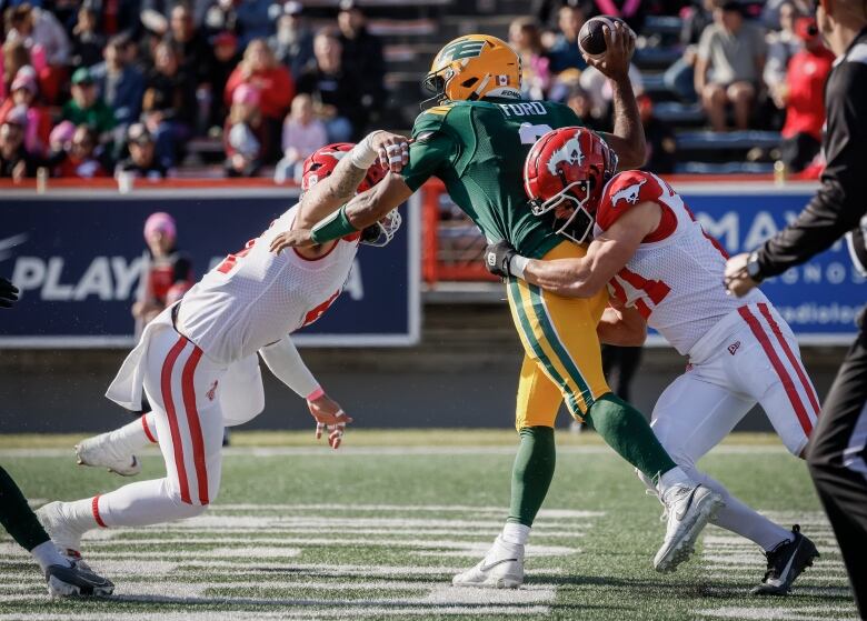 A football player in a green-and-yellow uniform is trying to throw the ball, while getting tackled by two opposing players in red-and-white uniforms.