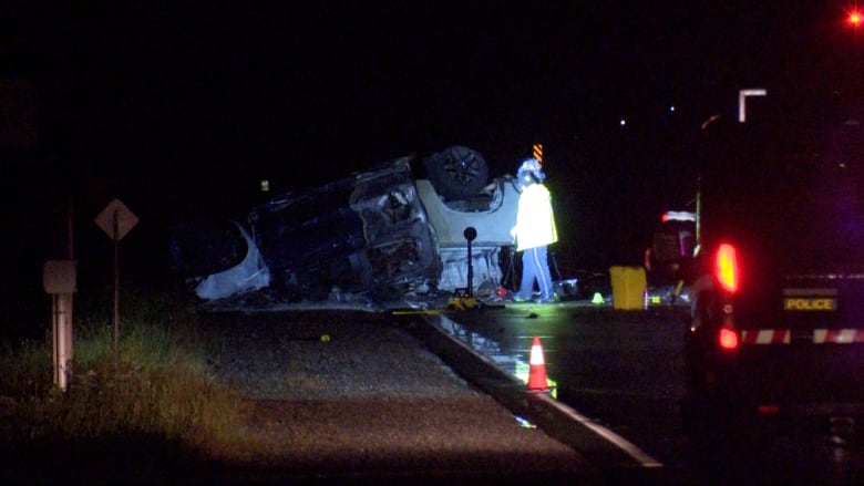 At night, a crumpled pickup truck lies on the side of a highway. A police vehicle is in the foreground. A person wearing reflective gear walks by the truck