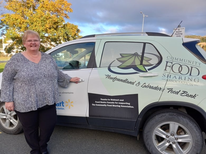 Woman with short blonde hair standing next to SUV with words Community Food Sharing Association on its side. There is a tree behind here that's leaves are green but it's also turning yellow and orange.