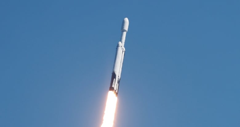 A white rocket is pictured mid-flight through blue skies, heading upwards with a jet of white flame tinged with red spitting out of the back of it. 