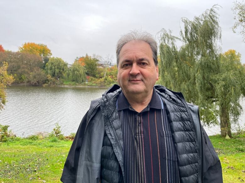 A man in a plaid shirt, wearing a light black jacket over top, poses for a picture outdoors. A river and weeping willow are seen behind him. 